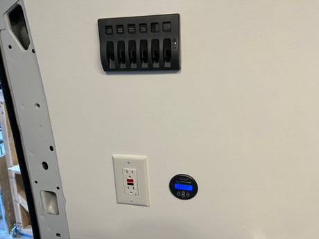 RV rental cabin interior featuring a control panel with switches, an electrical outlet, and a Victron energy battery monitor.