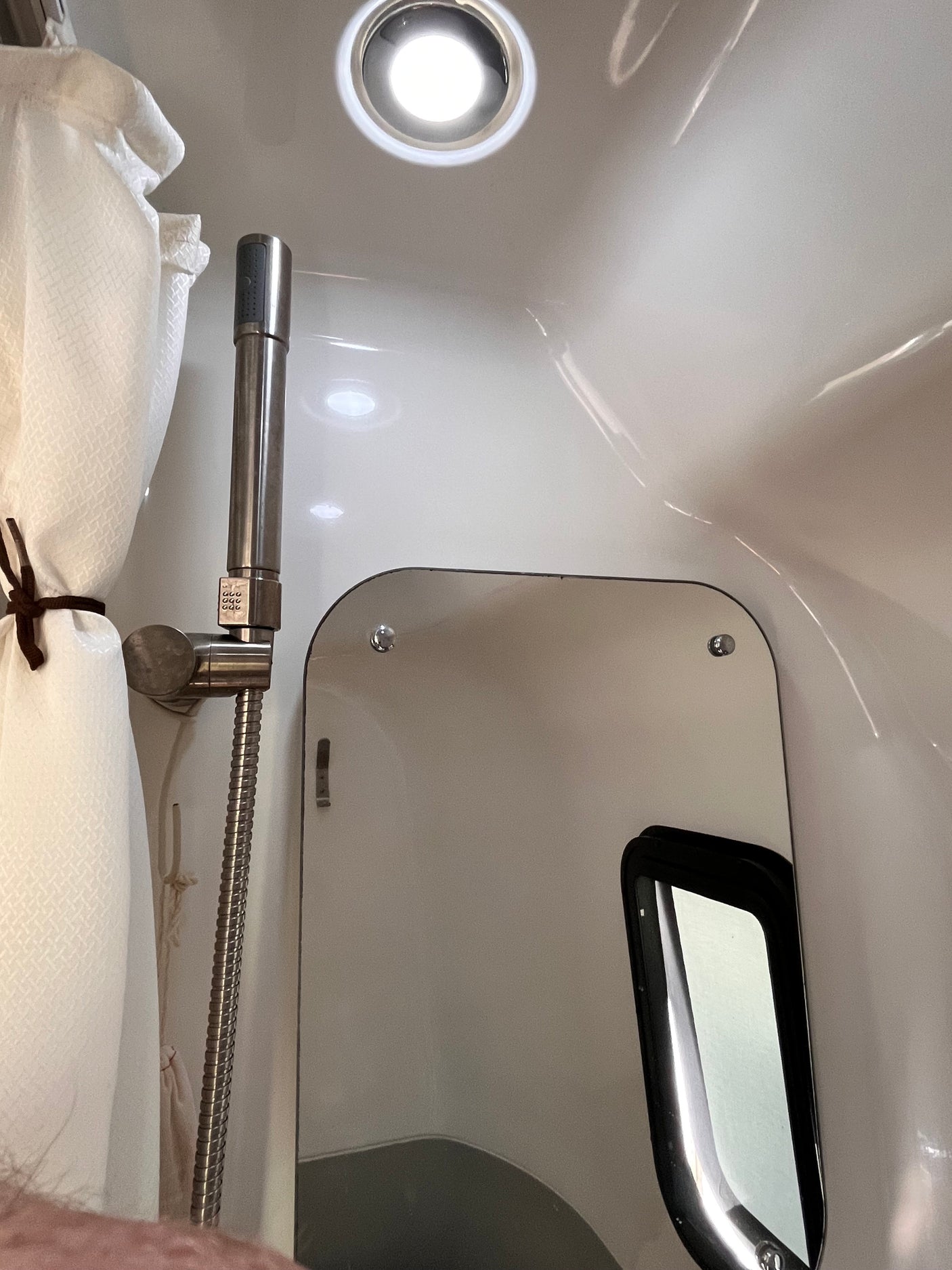 Inside a compact RV rental shower: chrome shower head, tied-back curtain, wall mirror, and ceiling light.