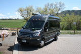 Dark grey Ford Transit van RV rental parked on pavers, with green fields and trees in the background.