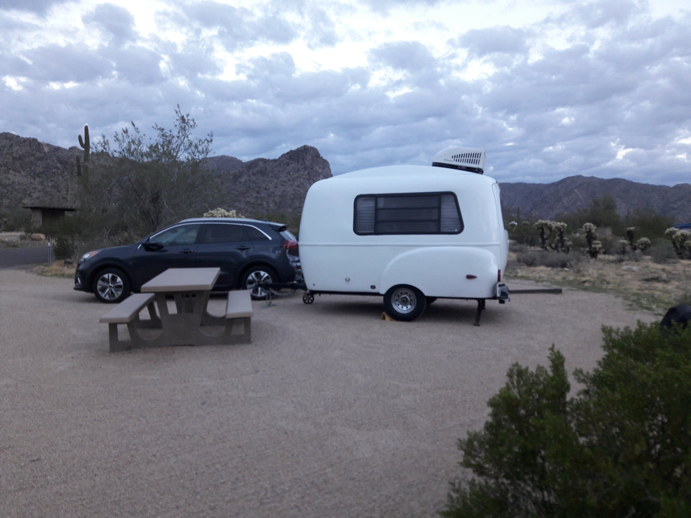A dark SUV tows a white RV rental teardrop camper at a desert campsite with a picnic table, mountains in background.