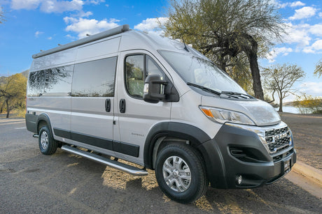 This silver Ram ProMaster RV rental is parked on an unpaved road near a scenic lake, ready for adventure.