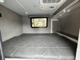 Interior of an RV rental showing a comfy bed, window, overhead AC, and storage for mobile living.