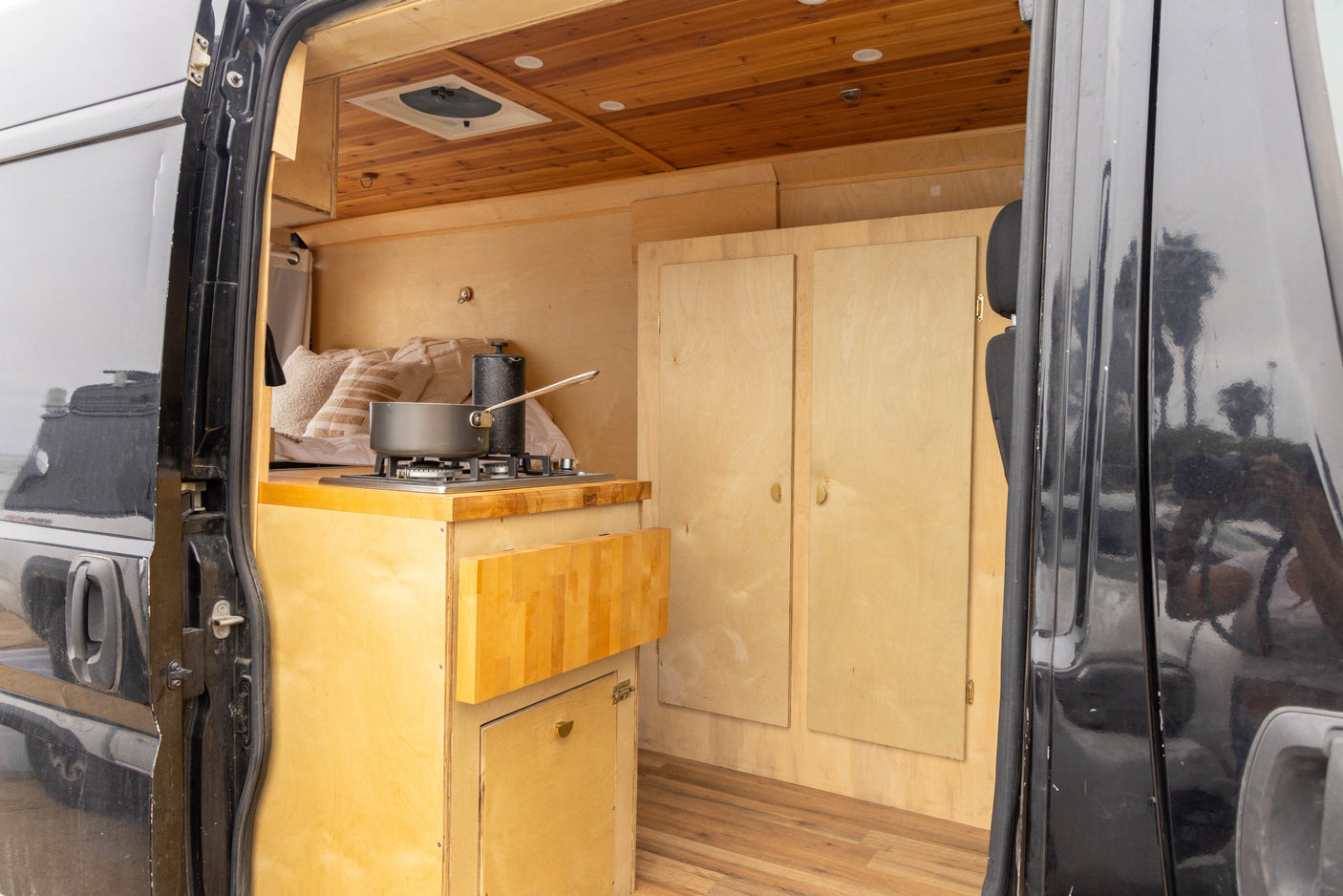 View into an rv rental showing its light wood interior with a compact kitchen stove, bed, and built-in storage.