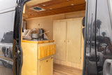 View into an rv rental showing its light wood interior with a compact kitchen stove, bed, and built-in storage.
