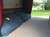 Inside an RV rental: a cozy bed with blue pillows and chocolate bars, red curtains opening to a grassy street with a toy car.