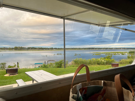 Scenic lake view from inside an RV rental, featuring a fire pit, picnic table, and calm water under a cloudy sky.