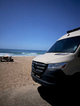 Night beach camping with an rv rental. A white camper van by the ocean under starry skies, picnic table nearby.
