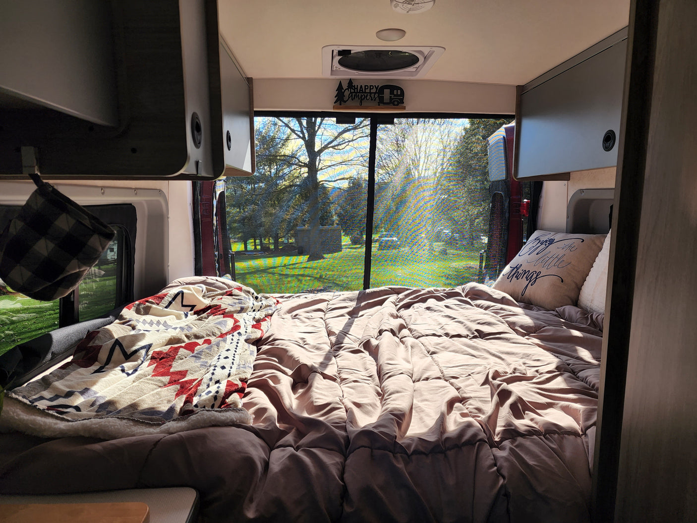 Bright RV rental interior showing a cozy bed and large window view of trees and sunlight. Perfect for travel!