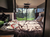 Bright RV rental interior showing a cozy bed and large window view of trees and sunlight. Perfect for travel!