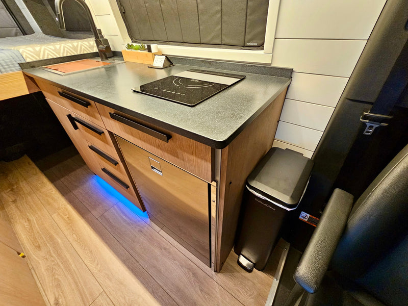 This modern RV rental kitchen features an induction cooktop, sink, fridge, storage, and blue under-cabinet lighting. — photo 19
