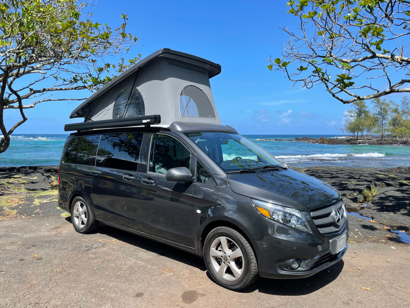 Dark grey pop-top camper van rv rental by the ocean, framed by trees. Ready for adventure on the coast.