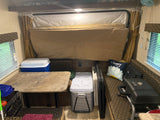 Inside a compact RV rental, featuring a comfortable bed, table, and seating with coolers and a grill.