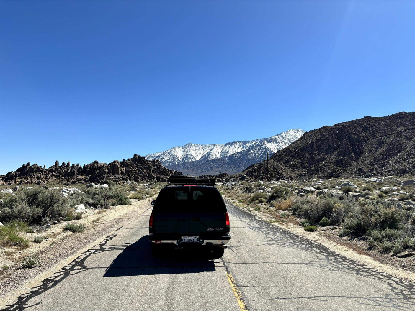 A dark SUV with a roof rack drives on a desert road towards majestic snow-capped mountains. Ideal for an RV rental trip.