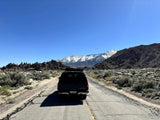 A dark SUV with a roof rack drives on a desert road towards majestic snow-capped mountains. Ideal for an RV rental trip.