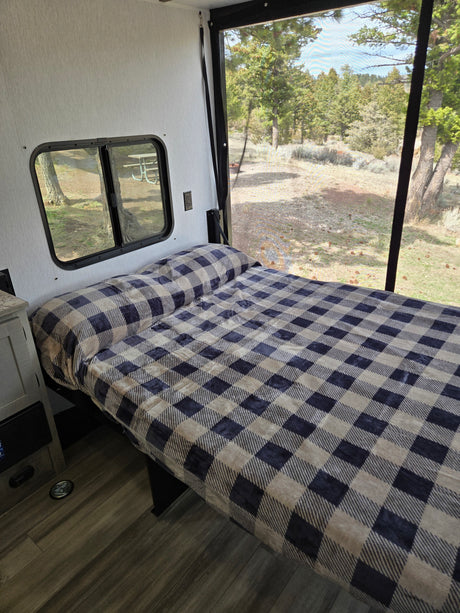 Cozy RV rental bed with plaid blanket & scenic nature views through window & screened door, perfect for camping.