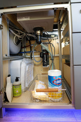 RV rental under-sink storage showing plumbing, water tanks, Frizzlife filter, and cleaning supplies like soap and Clorox wipes.