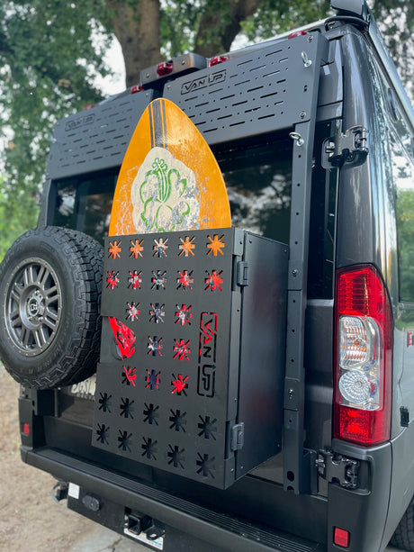 RV rental camper van with a black rear rack carrying a spare tire, storage box, & yellow surfboard.