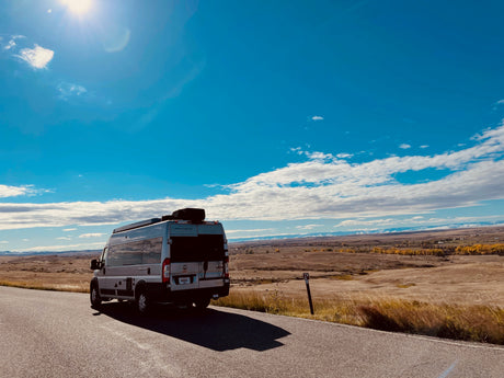 A modern RV rental parked on a scenic road, overlooking golden plains and distant snow-capped mountains under a bright sky.