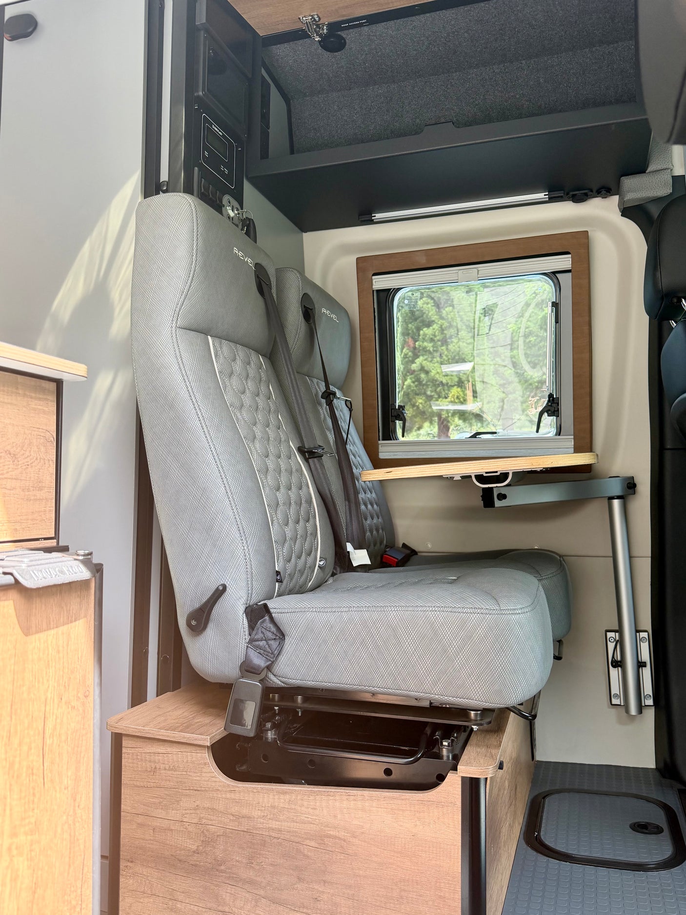 Inside this RV rental: two gray passenger seats with seatbelts, a retractable table, window, and control panel for camper van adventures.