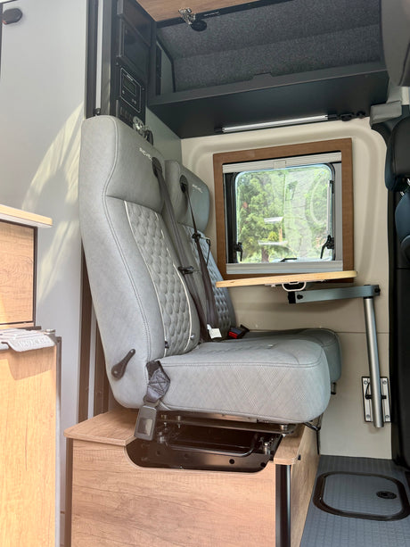 Inside this RV rental: two gray passenger seats with seatbelts, a retractable table, window, and control panel for camper van adventures.