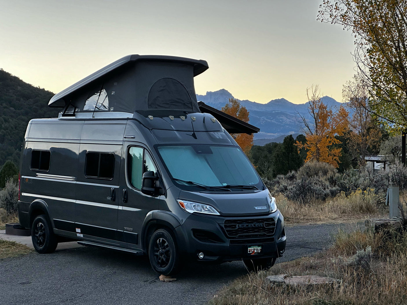 A dark gray pop-top camper van (RV rental) parked at a scenic campsite with mountains & autumn trees.
