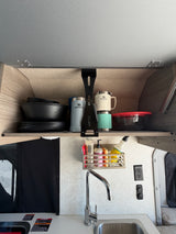 RV rental galley kitchen showing an open overhead cabinet with Stanley mugs, dishes, and a sink with faucet and cleaning supplies.