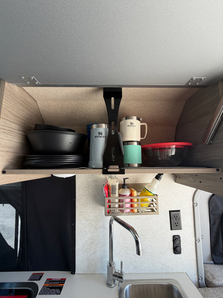 RV rental galley kitchen showing an open overhead cabinet with Stanley mugs, dishes, and a sink with faucet and cleaning supplies.