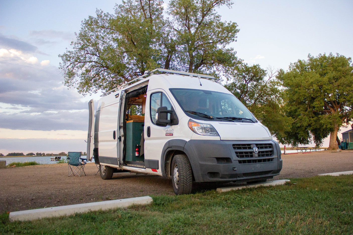 Enjoy the lakeside view from this spacious Ram ProMaster RV rental van, parked with its side door open and chair out.