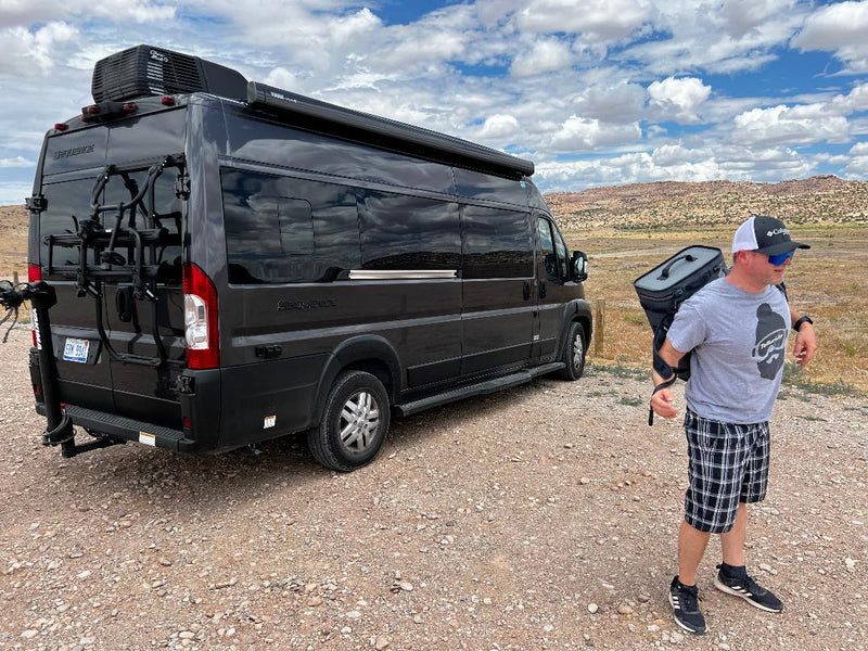  another RV in back.|Dark gray RV rental with a pop-top roof and extended awning parked in a sunny grassy field. Perfect for outdoor adventures.|Dark grey RV rental parked on a gravel road — photo 9