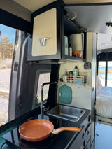 Inside an RV rental: compact kitchen with sink, induction cooktop, and a scenic window view to the beach.