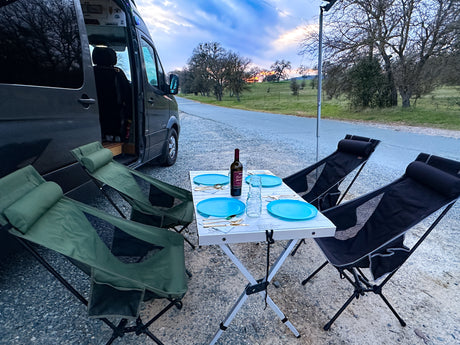 Scenic RV rental experience: Portable dining table with blue plates, wine, and camping chairs set beside a camper van at sunset.