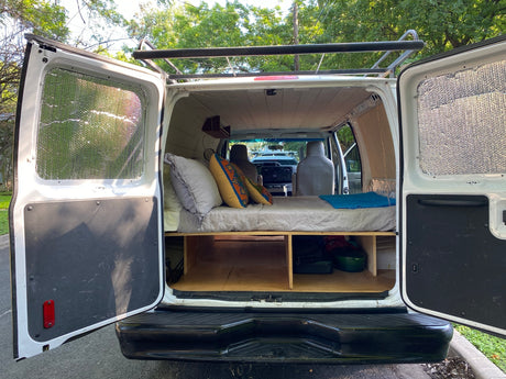 Rear view of a white van conversion, revealing a bed platform, storage, and insulated windows. Ideal for rv rental.
