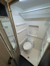View inside a compact RV rental bathroom, showing a white toilet, integrated shower area with shelves, and chrome rack.