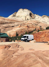 White RV rental van at the base of dramatic red & white sandstone mountains under a blue sky, ideal for scenic travels.