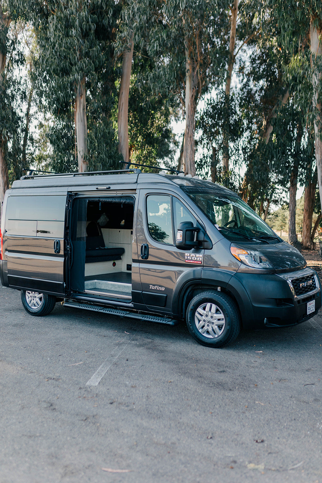 Dark gray Ram ProMaster camper van for rv rental, side door open showing cozy interior. Tall trees in background.