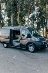 Dark gray Ram ProMaster camper van for rv rental, side door open showing cozy interior. Tall trees in background.