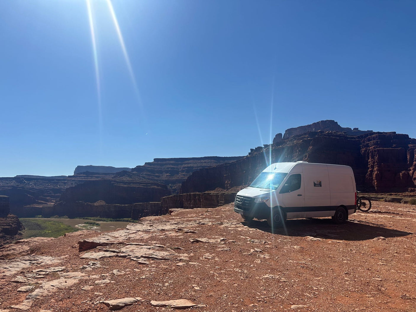 This RV rental, a white campervan, offers epic views from a red rock canyon overlooking a river under a bright sky.