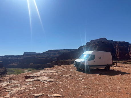 This RV rental, a white campervan, offers epic views from a red rock canyon overlooking a river under a bright sky.
