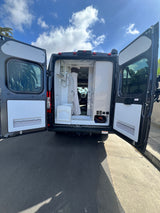 Open rear doors of a dark grey RV rental reveal a compact interior with a shower, toilet, and storage. Parked on asphalt.