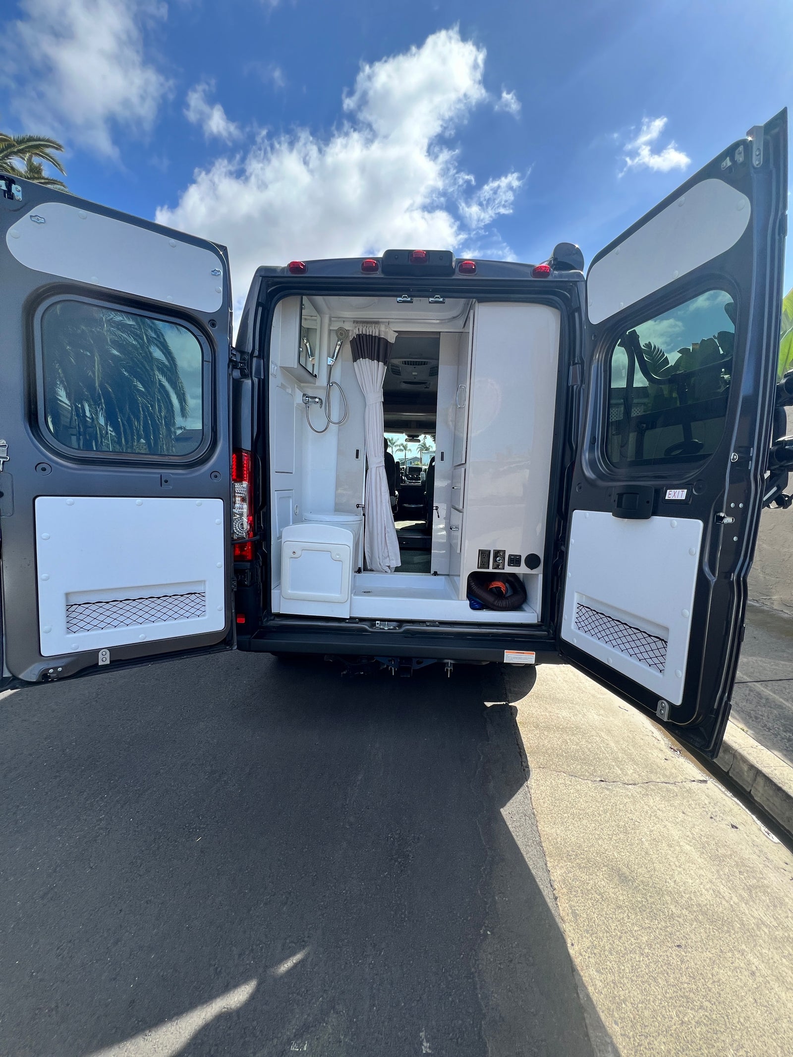 Open rear doors of a dark grey RV rental reveal a compact interior with a shower, toilet, and storage. Parked on asphalt.