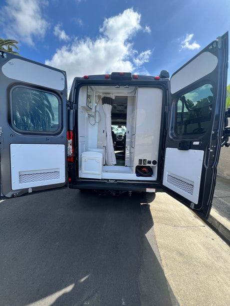 Open rear doors of a dark grey RV rental reveal a compact interior with a shower, toilet, and storage. Parked on asphalt.