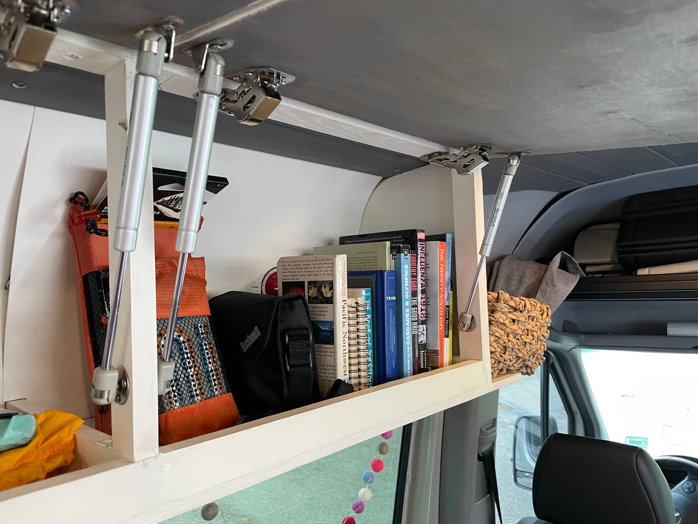 Open overhead storage in an RV rental, neatly filled with books, a Bushnell bag, and travel essentials for the road.