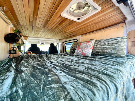 RV rental camper van interior: A cozy bed with green crushed velvet bedding, wooden slatted ceiling, and roof vent fan.