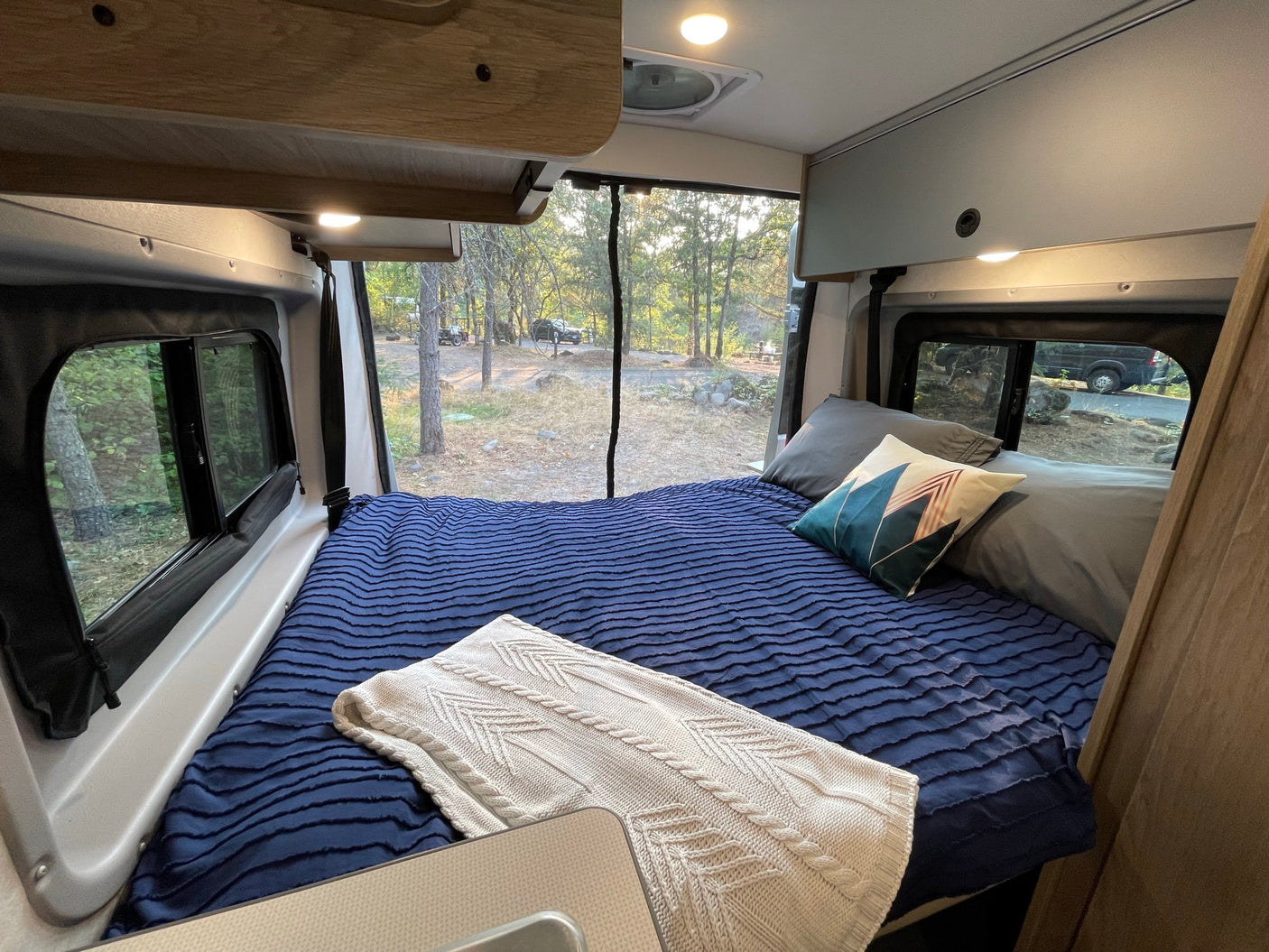 An inviting RV rental bedroom with a blue bed and knitted throw, overlooking a peaceful, wooded campsite.