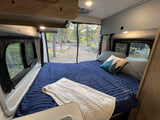 An inviting RV rental bedroom with a blue bed and knitted throw, overlooking a peaceful, wooded campsite.