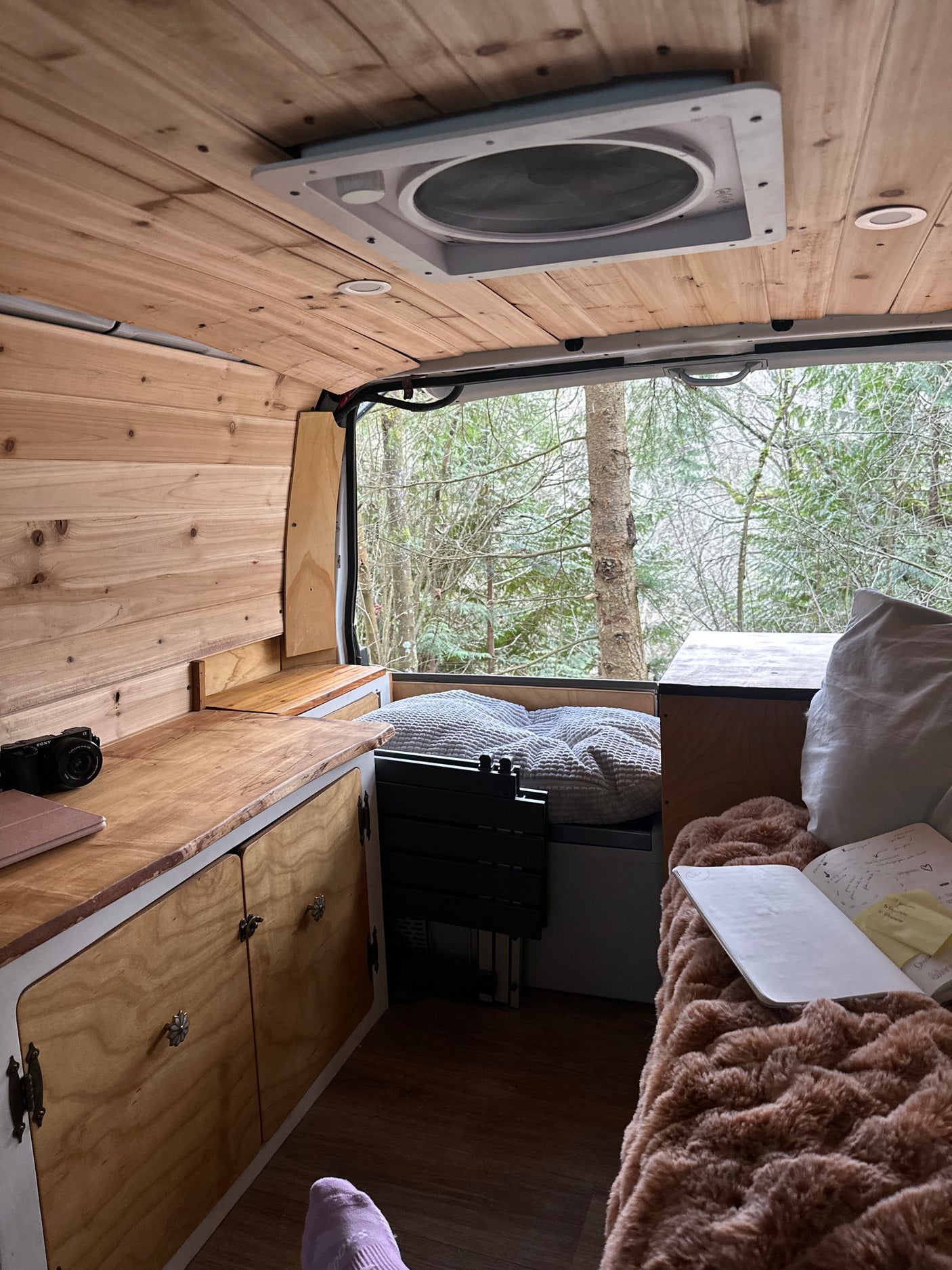 Cozy RV rental interior featuring custom wood, plush bed, work area, and a tranquil forest view. Ideal for travel.