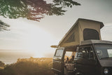 This RV rental offers stunning ocean sunset views. A dark pop-top camper van is parked, with two people relaxing inside.