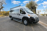 This silver camper van RV rental awaits its next journey, parked on a paved road in a sunny, tree-lined park.