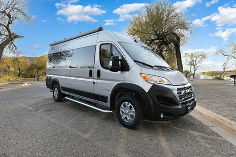 This silver camper van RV rental awaits its next journey, parked on a paved road in a sunny, tree-lined park.