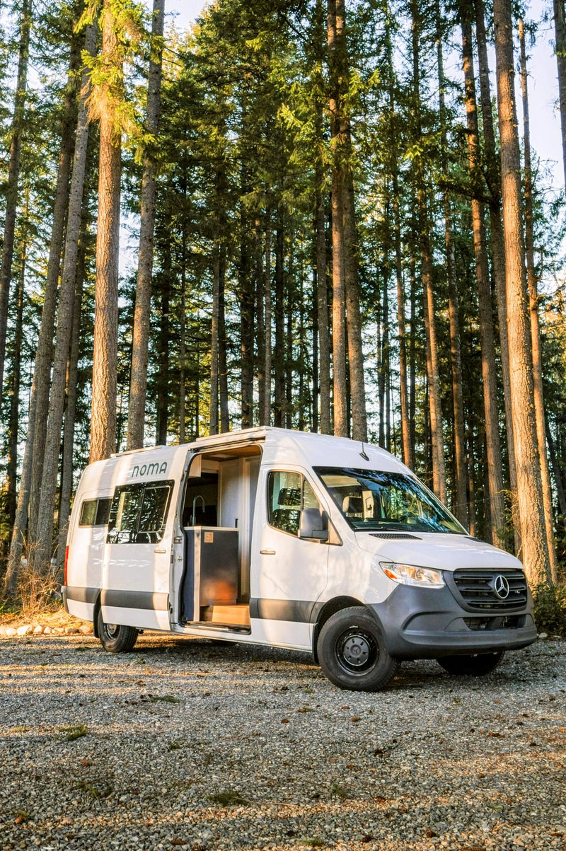 A white Mercedes Sprinter RV rental camper van with its side door open, parked on gravel amidst tall forest trees. — photo 17